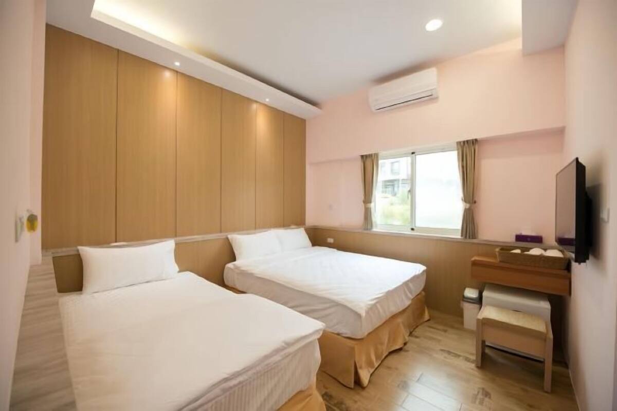 A hotel room with two single beds, a wooden headboard, a small desk with a chair, a window with curtains, and an air conditioning unit.