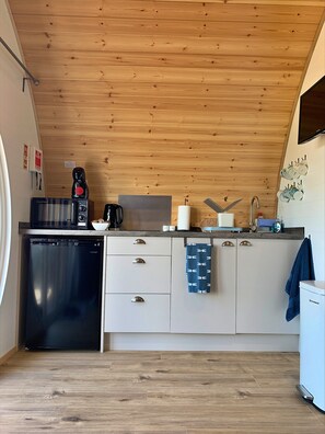 A compact kitchen with wooden ceiling, white cabinets, and a black dishwasher. - Glamping Pods in Yorkshire Near York & Coast (York)
