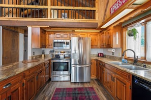 A kitchen with wooden cabinets, stainless steel appliances, a granite countertop, and a red plaid rug. - Cozy Cabin w hot tub & wood-burning fireplace (Whitefish)