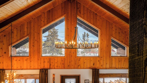 Interno di una baita in legno con ampie finestre, un lampadario e vista sull'esterno.