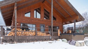 Una baita di legno con veranda coperta, ampie finestre e una pila di legna davanti.