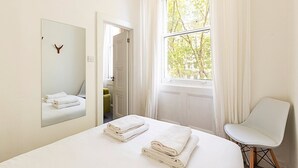 A hotel room with a bed, a chair, a mirror, and a window with a view of trees. - Elegant Knightsbridge Flat Near Harrods (London)