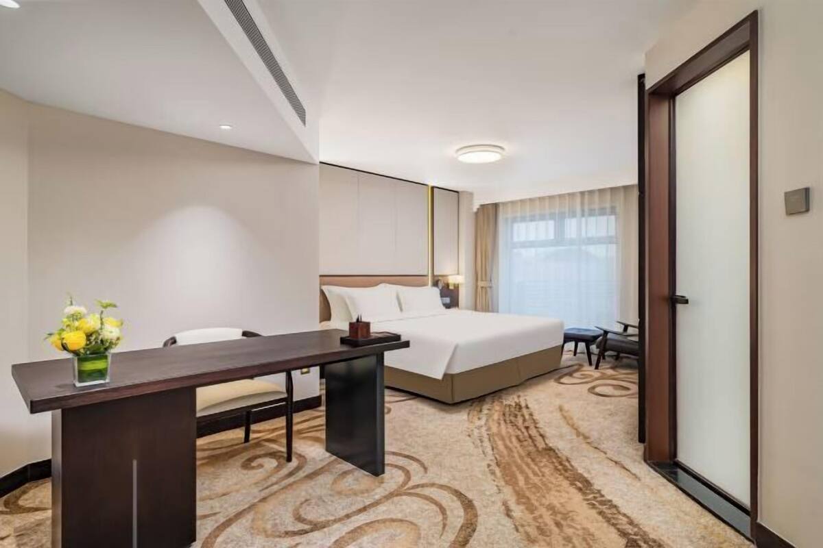 Habitación de hotel con cama, escritorio, silla, una mesita con un florero y un ventanal con cortinas.