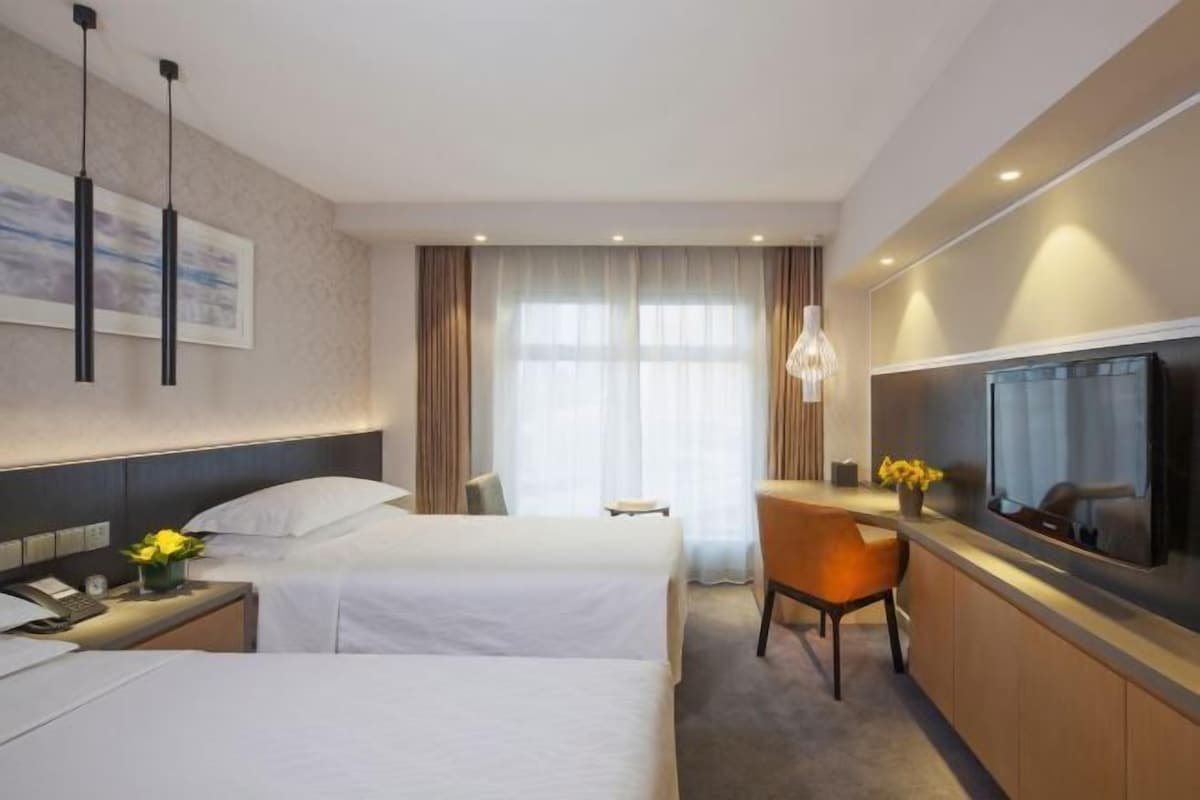 A hotel room with two beds, a desk with a chair, a TV, and a large window with curtains.