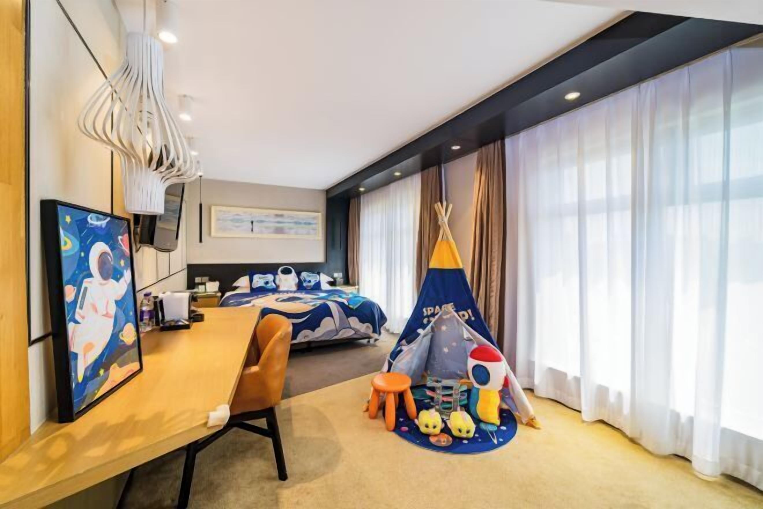A hotel room with a bed, a desk, a chair, a window with curtains, a painting, and a children's play area with a teepee and toys.