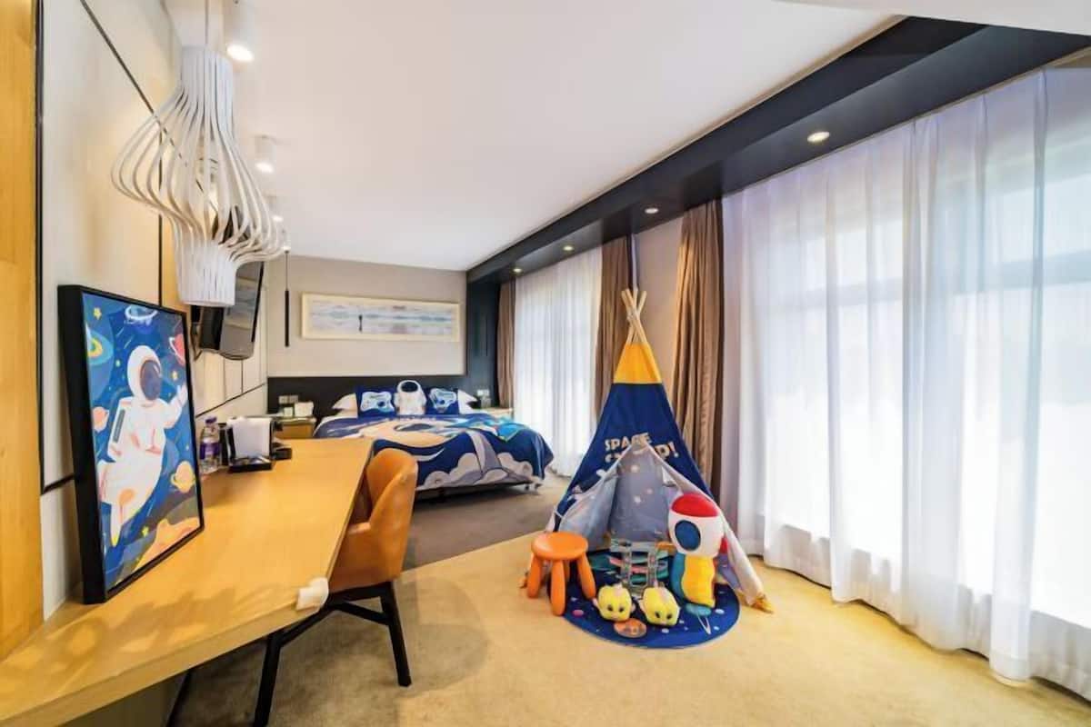 Habitación de hotel con cama, escritorio, silla, ventana con cortinas, un cuadro y una zona de juegos infantil con un tipi y juguetes.