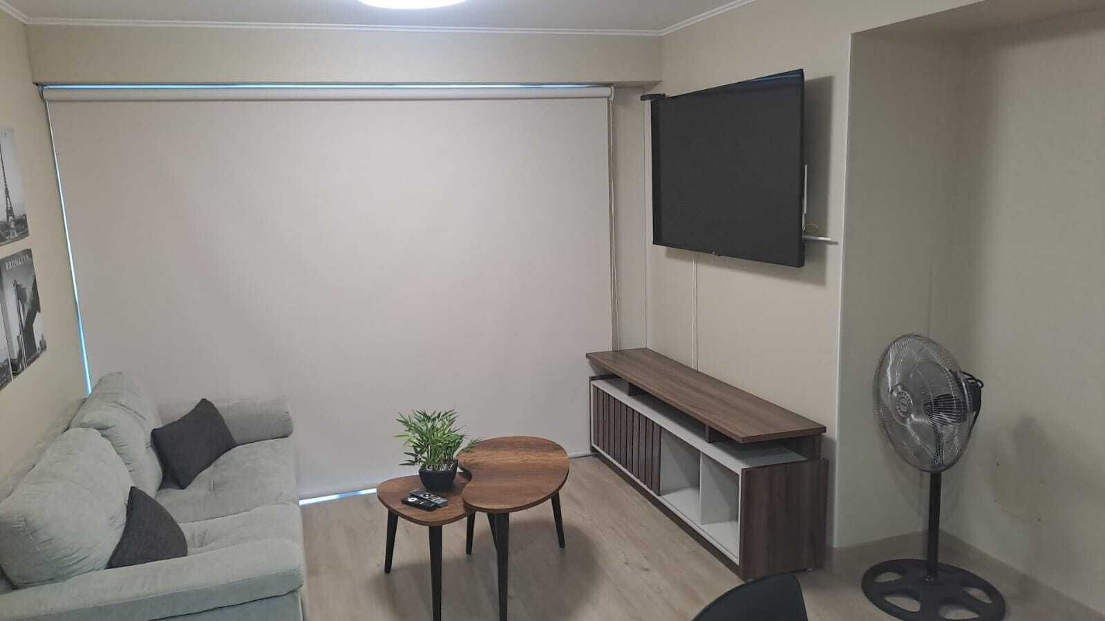Een woonkamer met een bank, een bijzettafeltje, een kleine plant, een aan de muur bevestigde televisie en een staande ventilator.