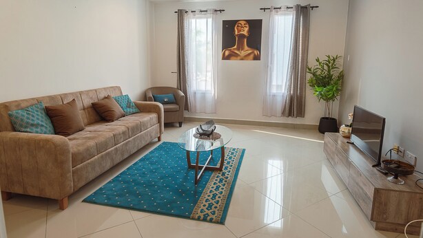 A modern living room with a sofa, a glass coffee table, a TV, and a large window with curtains. - Modern 3-bedroom townhome with a pool, AC and Wi-Fi in vibrant East Legon (Accra)