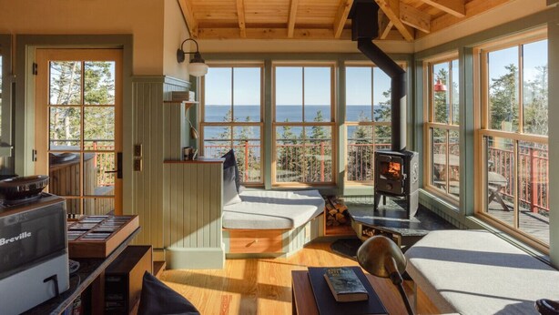 Un intérieur de chalet chaleureux avec un poêle à bois, de grandes fenêtres donnant sur l’océan et un appareil de la marque Breville placé sur la gauche.