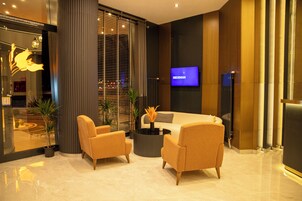 Um lobby moderno com um sofá, duas poltronas, uma mesa de centro e uma tela de TV exibindo 'DEDEMAN'.
