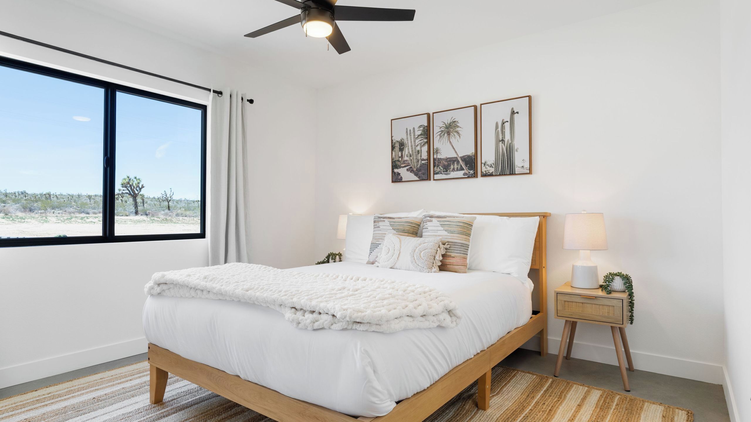 A bedroom with a large window, a ceiling fan, a bed with white bedding, a nightstand with a lamp, and framed desert-themed artwork on the wall.