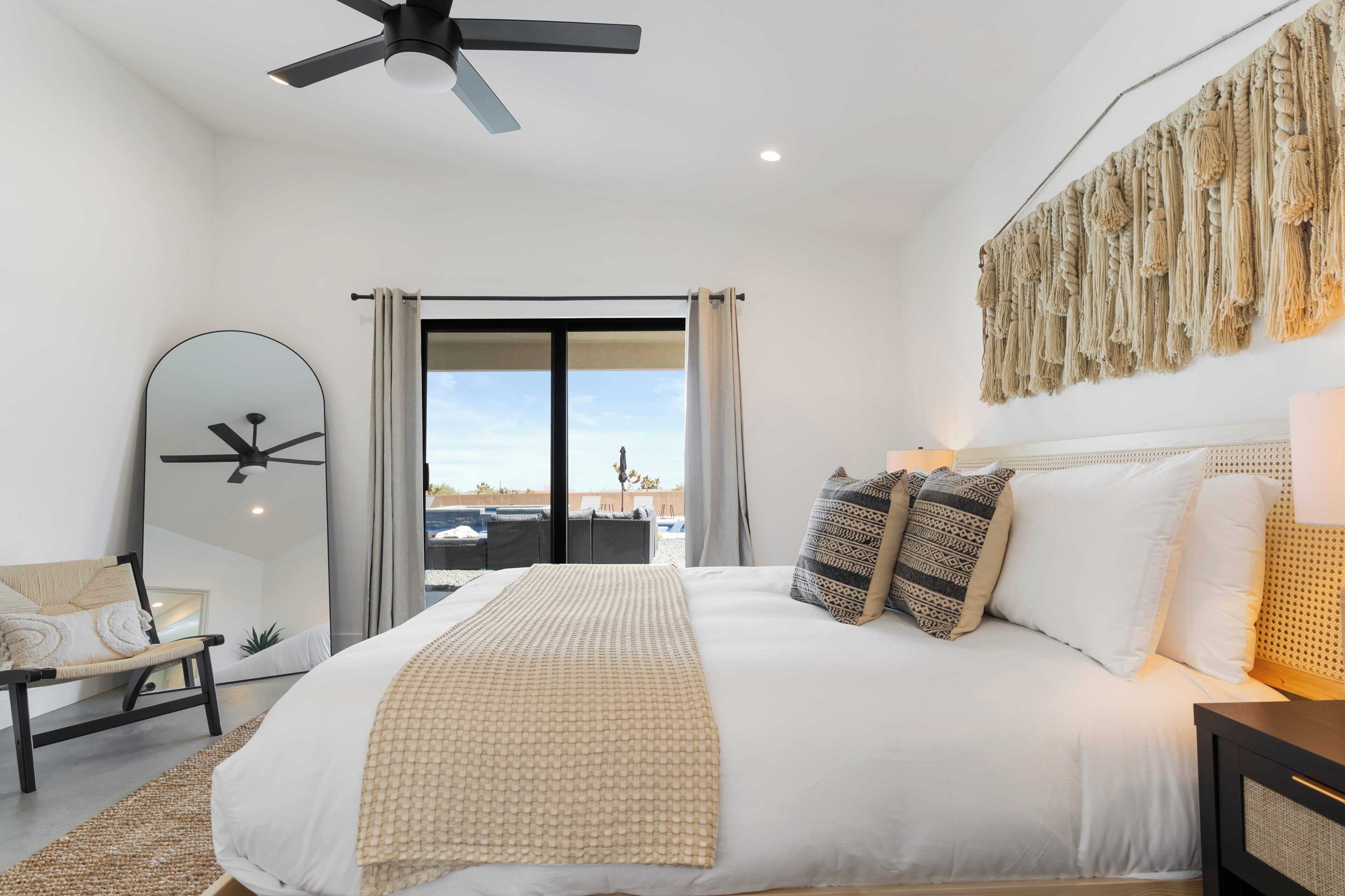 A modern bedroom with a large bed, a ceiling fan, a large mirror, and a view of the outdoors.