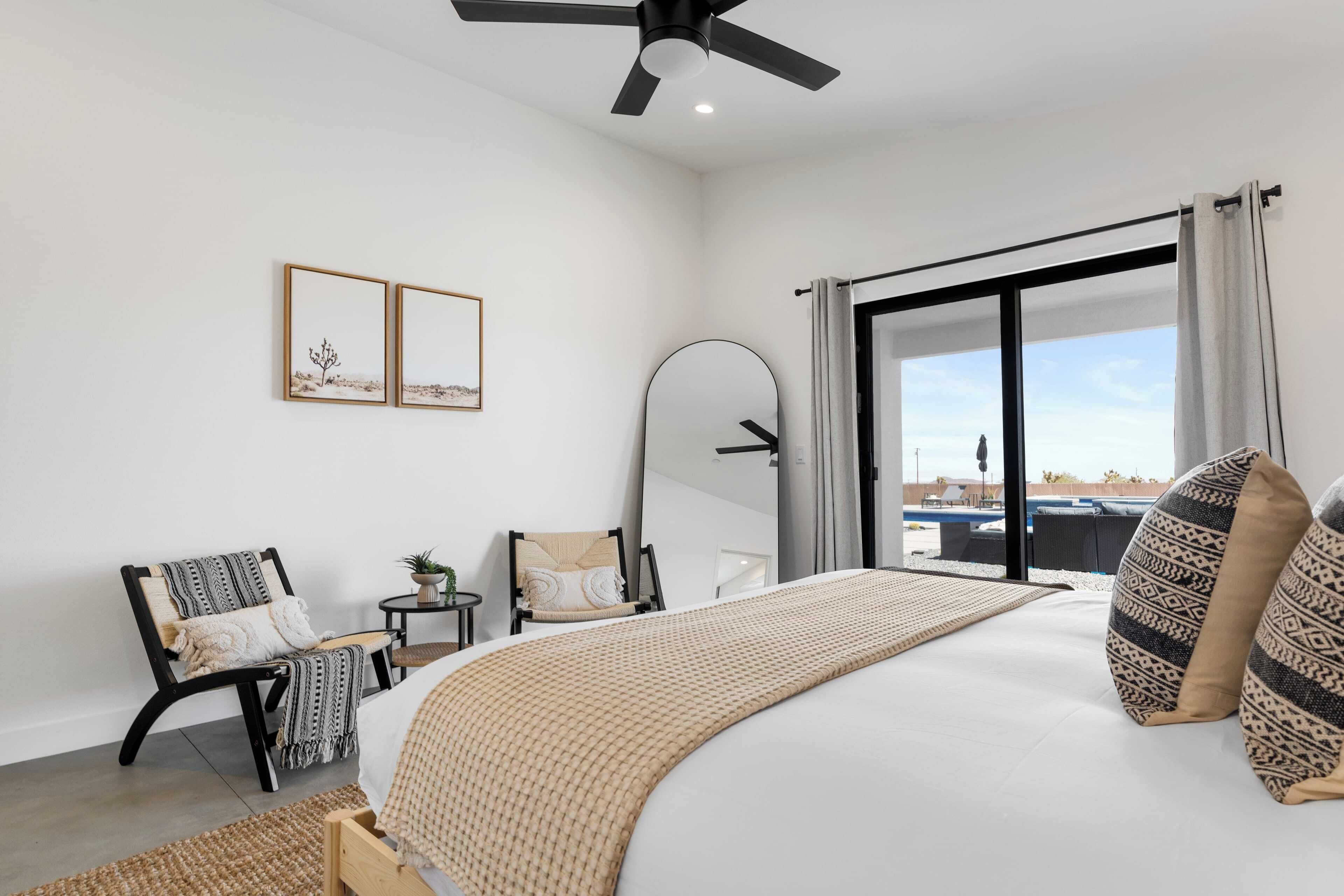 Una camera da letto moderna con un letto grande, una ventola a soffitto, una scrivania con una sedia, un tavolino con una pianta e vista sull'esterno tramite una porta scorrevole.