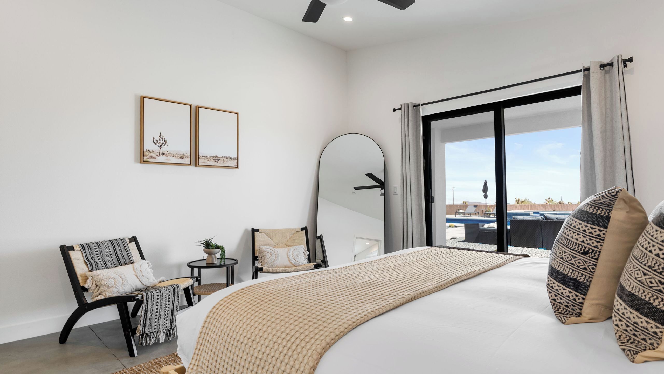 Een slaapkamer met een bed, een stoel, een klein tafeltje met een plant, een spiegel en uitzicht op een buitenruimte.