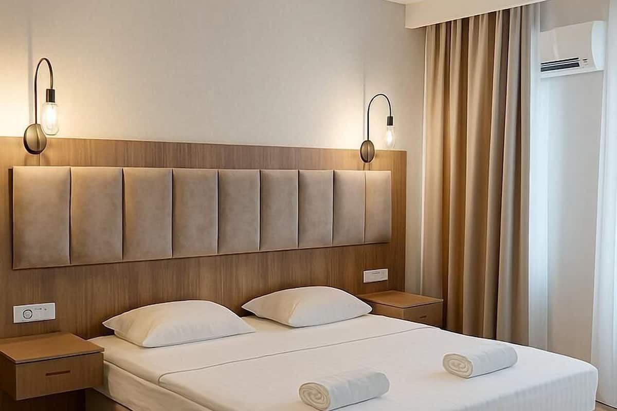 A modern hotel room with a large bed, wooden headboard, bedside tables, and a wall-mounted air conditioner.