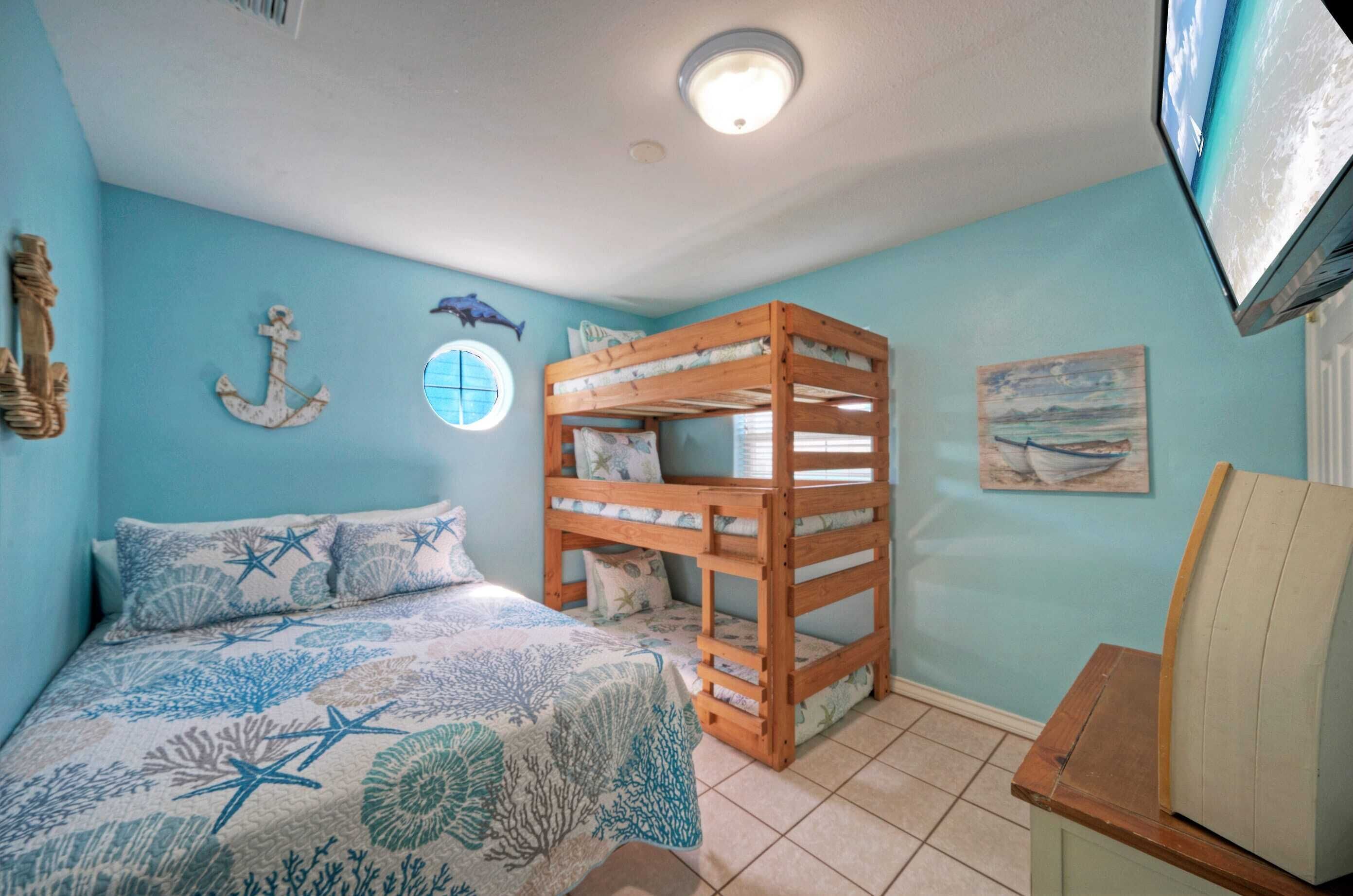Une chambre avec un lit superposé, un lit recouvert d’une couette à motifs d’étoiles de mer, une fenêtre ronde et un tableau représentant un bateau accroché au mur.