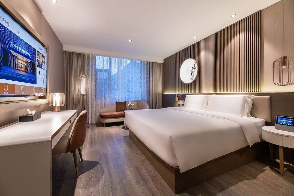 A modern hotel room with a large bed, a desk, a chair, and a TV displaying hotel information.