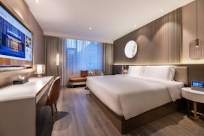 A modern hotel room with a large bed, a desk, a chair, and a TV displaying hotel information.