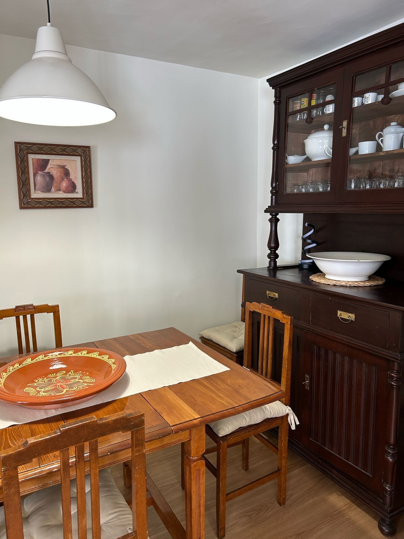 A dining area with a wooden table, chairs, a white pendant light, a cabinet with glass doors, and a framed picture on the wall.