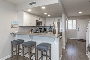 Una cocina moderna con armarios blancos, electrodomésticos de acero inoxidable y encimera gris. Hay tres taburetes de bar y una ventana con persianas.