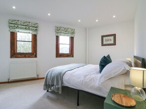 Ein Schlafzimmer mit einem Bett, zwei Fenstern mit Vorhängen, einem Heizkörper, einem gerahmten Bild an der Wand und einem Nachttisch mit Lampe und einem Holztablett.