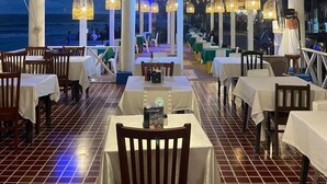 Un área de comedor al aire libre con mesas y sillas, un ventilador de techo decorativo grande y vista al océano.