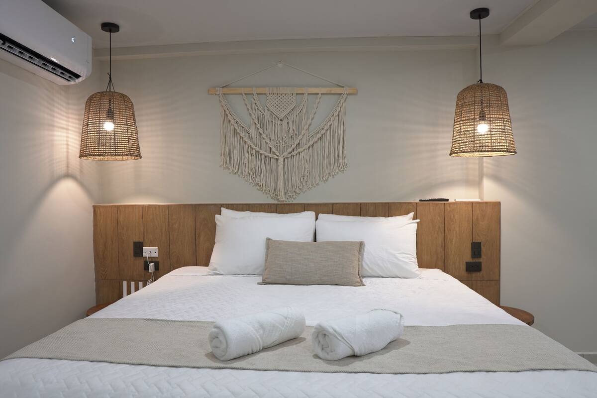 A neatly made bed with white linens, a wooden headboard, and two hanging macrame wall hangings.