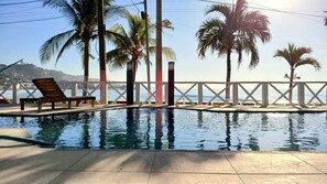 A swimming pool with a wooden deck and lounge chairs, surrounded by palm trees and a view of the ocean. - Litos Hotel y Restaurante (La Libertad)