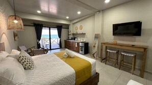 Habitación de hotel con cama, escritorio con televisor, balcón con vistas y kitchenette.