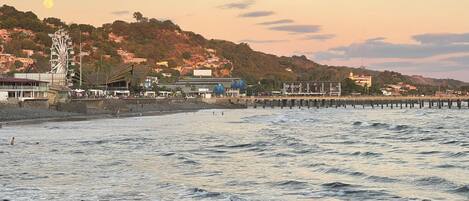 A coastal scene with a rocky beach, a pier extending into the water, and buildings on a hillside in the background.