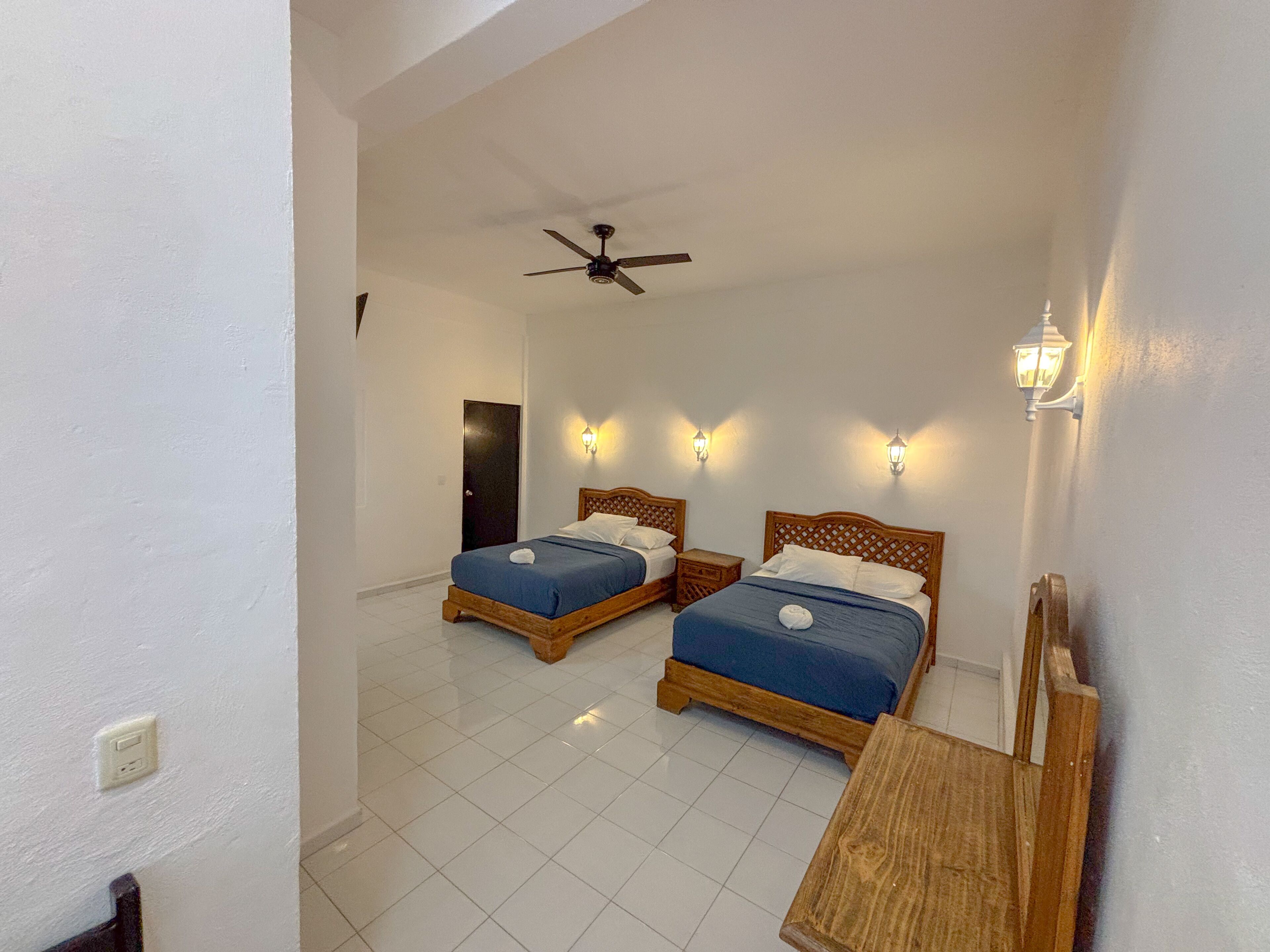 A hotel room with two beds, a ceiling fan, and wall-mounted lights.