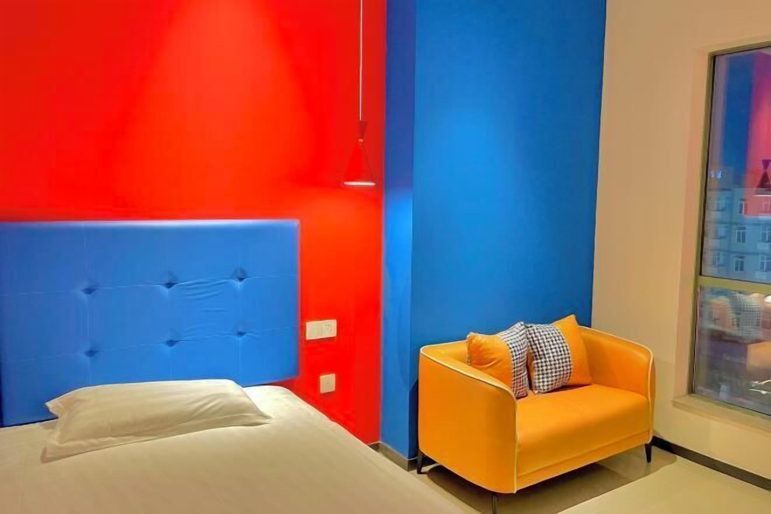 A room with a bed, a blue headboard, an orange armchair, and a window with a city view.