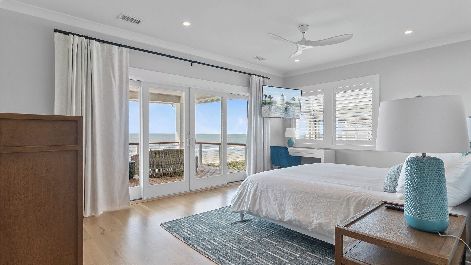 Een slaapkamer met een bed, een bureau, een stoel, een lamp en uitzicht op het strand.