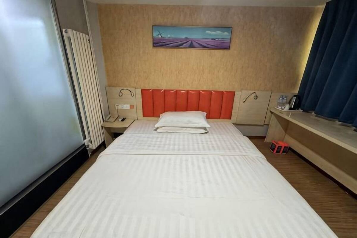 Ein Hotelzimmer mit Bett, Kopfteil, Nachttisch, Vorhängen an einem Fenster und einem Bild an der Wand.