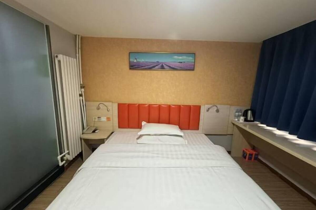 Ein Hotelzimmer mit Bett, Kopfstütze, einem Gemälde an der Wand, einem Fenster mit Vorhängen und einem Heizkörper.