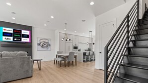 A modern living room with a grey sofa, a dining table with chairs, a kitchen area, and a staircase.
