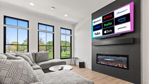 A modern living room with a sectional sofa, a flat-screen TV mounted on the wall, a fireplace, and large windows offering a view of the outdoors.