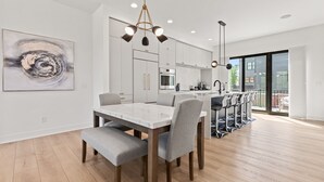 A modern kitchen with white cabinets, a central island, and a dining area with a marble table and grey upholstered chairs.