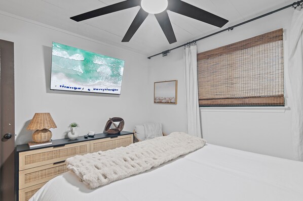 A bedroom with a bed, a ceiling fan, a wall-mounted TV, a bedside table with a lamp, a small plant, and a window with blinds.