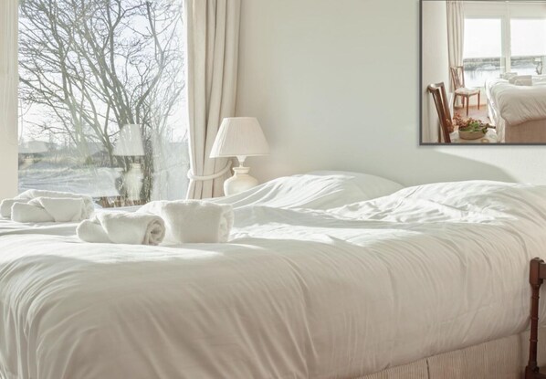 Un lit bien fait, avec du linge de lit et des serviettes blanches, une lampe de chevet et une fenêtre donnant sur des arbres et une étendue d’eau.