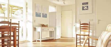 Una habitación luminosa con suelo de madera, un escritorio blanco, sillas de mimbre y una mesa de centro de vidrio con objetos decorativos.