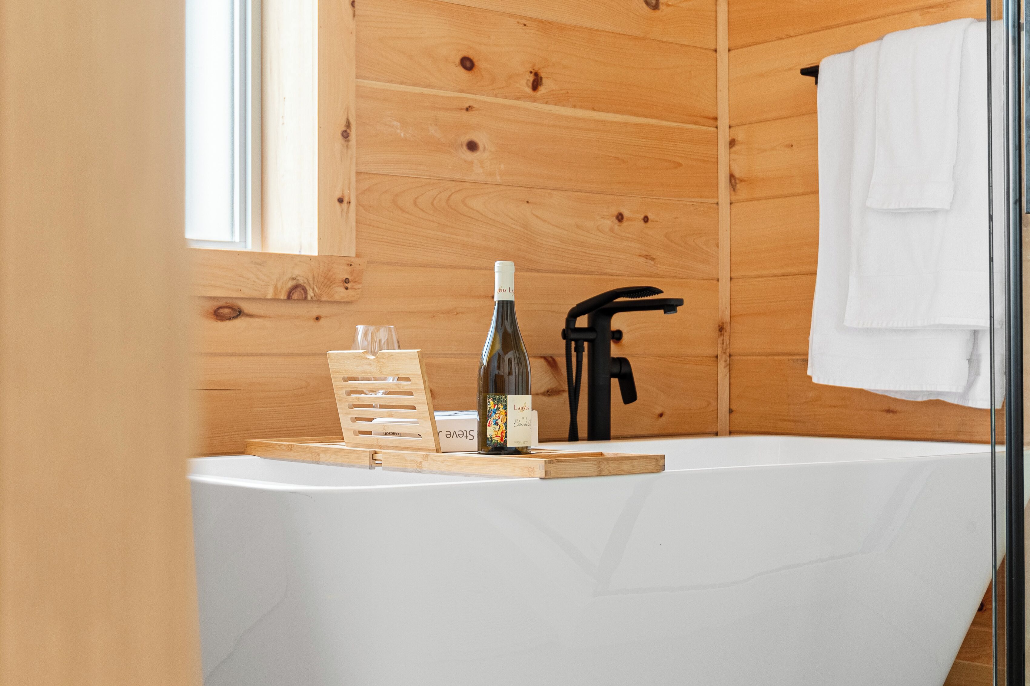 Une salle de bain rustique, avec des murs en bois, une baignoire blanche, une bouteille de vin et un porte-serviettes.