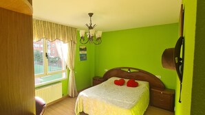 Una habitación con cama, ventana con cortinas, radiador, cabecero de madera, mesita de noche y una lámpara fijada a la pared.