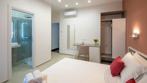 A modern hotel room with a glass-enclosed shower, a desk with a chair, a bed with red pillows, and a wall-mounted air conditioning unit. - RossyPalace - Amalficoast Suites (Agerola Bomerano)