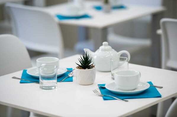 Une table à manger dressée avec de l’assiette blanche, des tasses et une petite plante en pot.