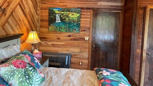 A wooden-paneled room with a bed, a nightstand, a lamp, and a painting of a waterfall. - Magic Skies Top Floor Waterfall Botanical Room (Hilo)