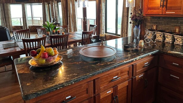 A spacious kitchen with wooden cabinets, a granite countertop, and a dining area with a table and chairs. - Magic Skies Top Floor Waterfall Botanical Room (Hilo)