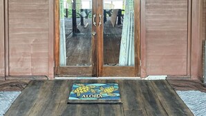 A wooden door with a ship's wheel decoration above it, a glass door with two handles, and a mat with 'ALOHA' and turtle illustrations at the entrance. - Magic Skies Top Floor Waterfall Botanical Room (Hilo)