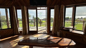 A wooden interior with a large table, large windows, and a view of the outdoors. - Magic Skies Surfs Up Room (Hilo)