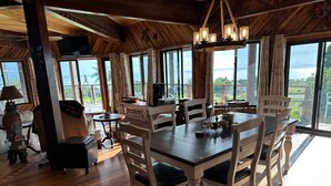 A spacious dining area with a large wooden table and chairs, large windows offering a view of the outdoors, and a cozy seating area with a lamp and decorative items. - Magic Skies Surfs Up Room (Hilo)